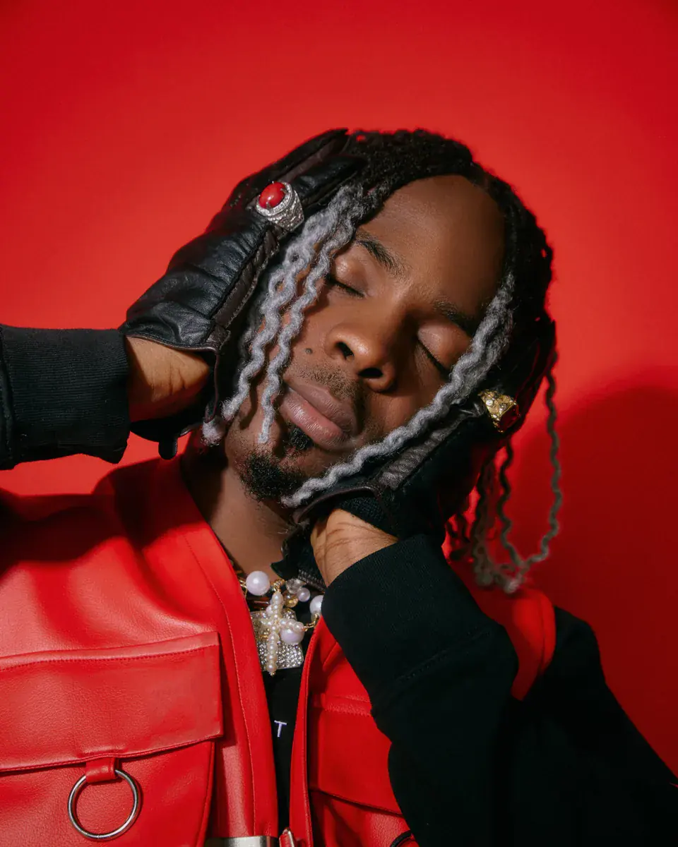 Young Jonn with silver-streaked locs, eyes closed, holding his head in gloved hands against a red background