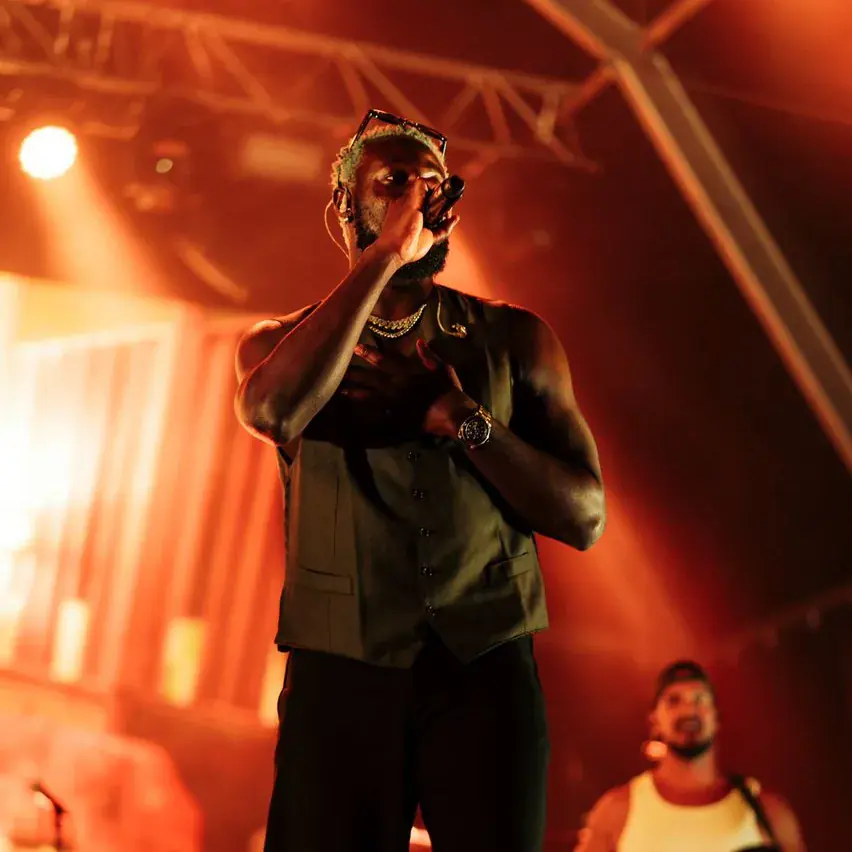 Kel-P performing on a stage lit orange, holding a microphone to his mouth, with a bassist behind him