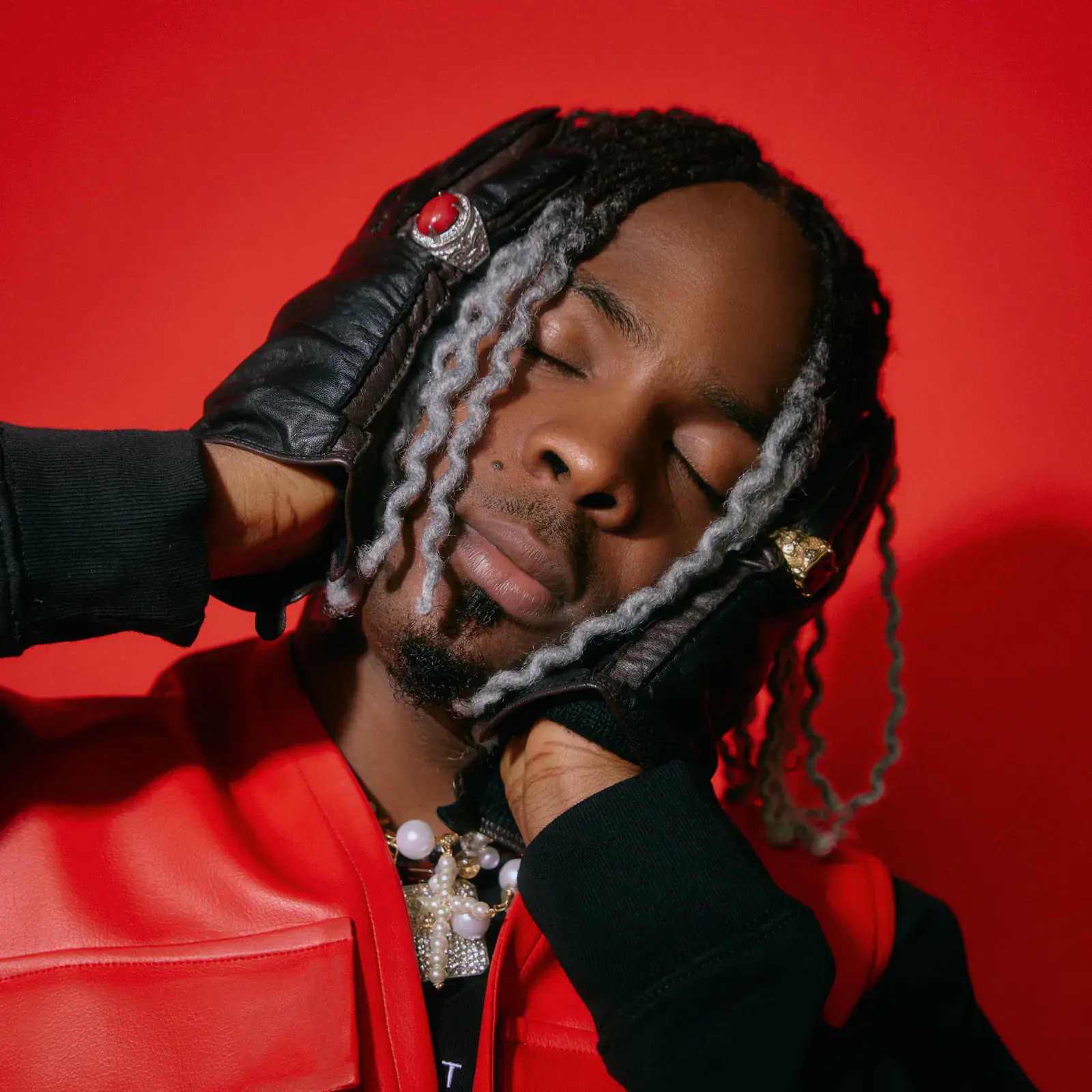 Young Jonn with silver-streaked locs, eyes closed, holding his head in gloved hands against a red background