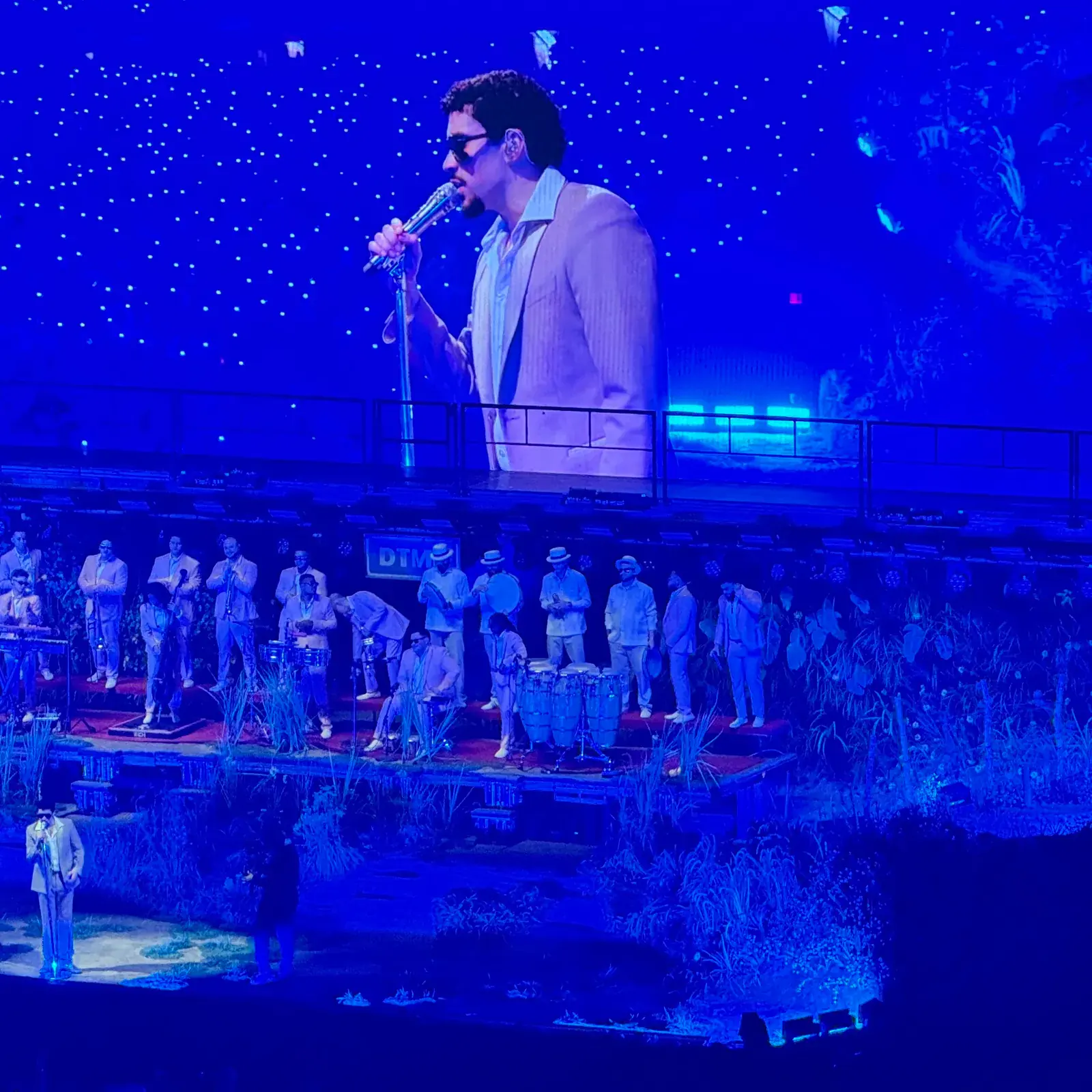 Bad Bunny sings below a giant screen showing his performance, with musicians behind him in blue stage lighting