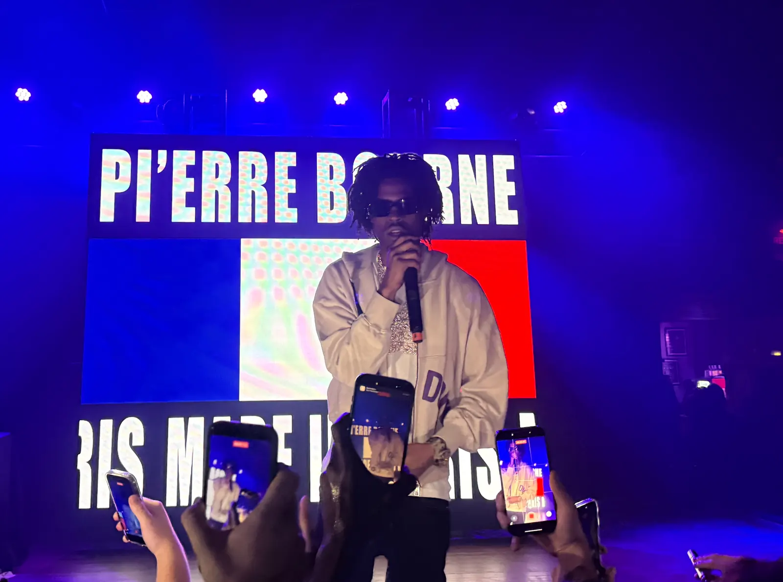 Pi’erre Bourne holds a microphone in front of a bright stage screen while audience members film on phones