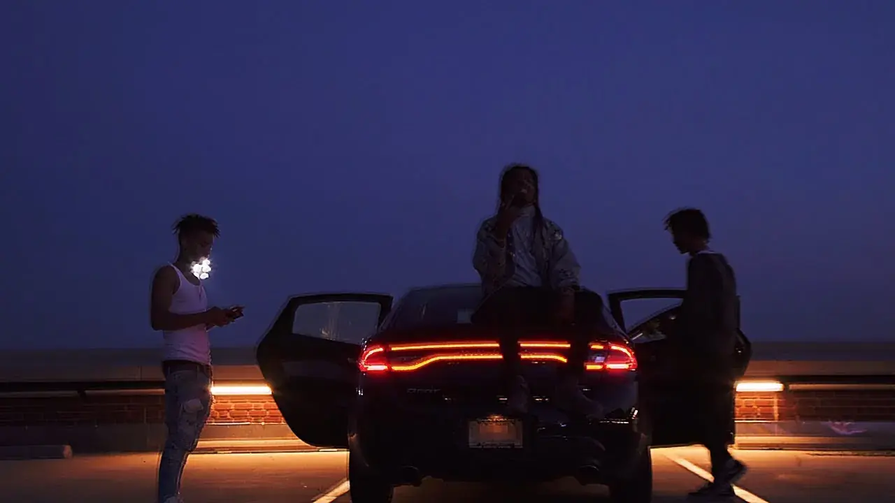 Three men stand around a black car with glowing red rear lights at dusk