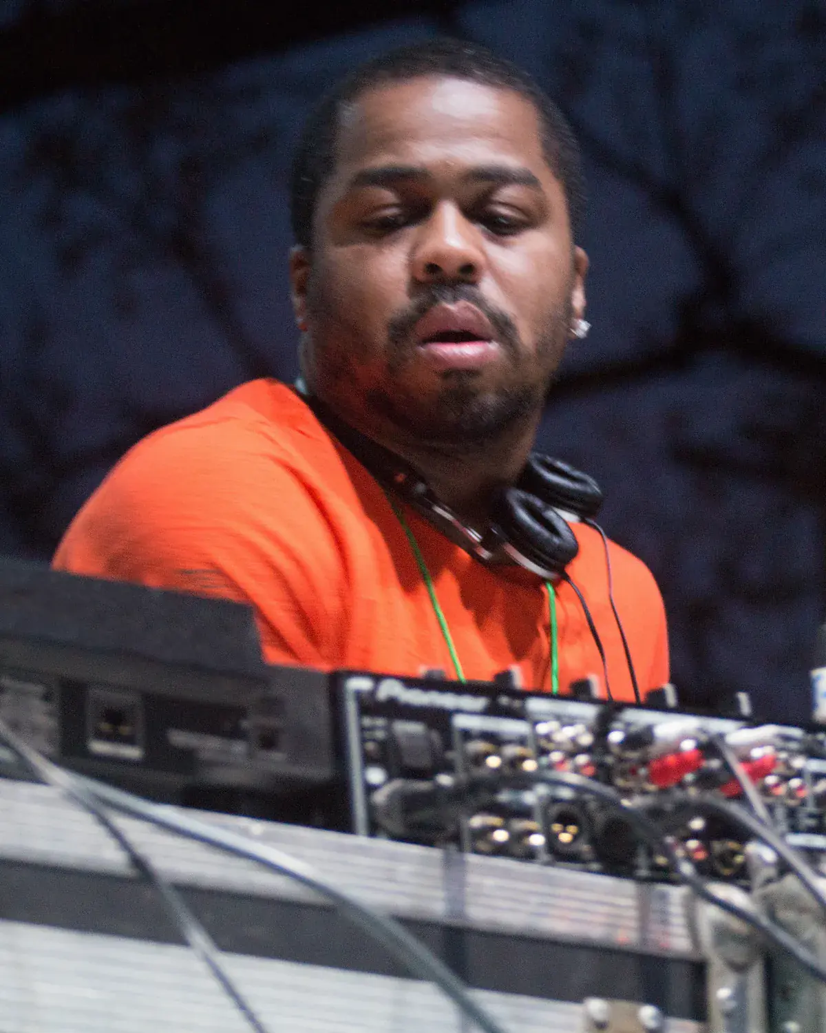 Just Blaze in an orange T-shirt and headphones behind a DJ mixer at an outdoor festival
