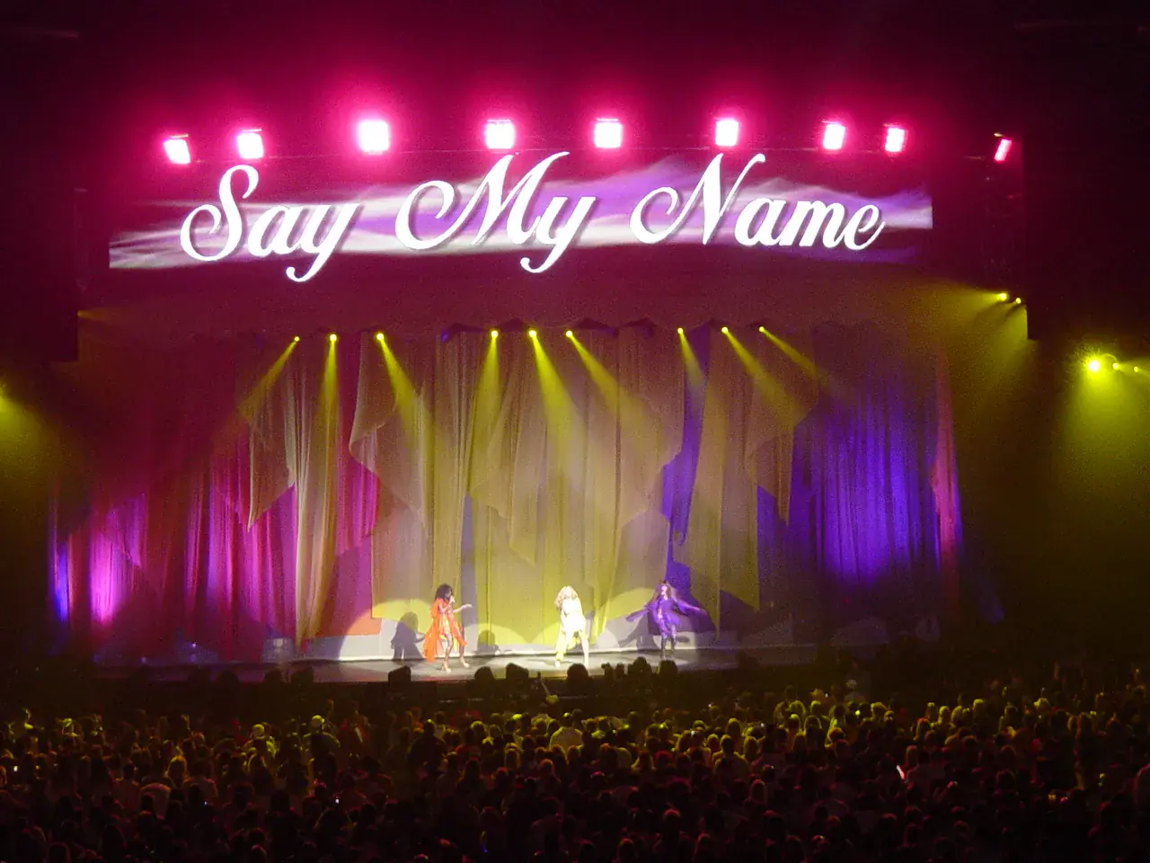 A packed arena facing a stage lit pink and gold, with three performers under a giant sign reading Say My Name