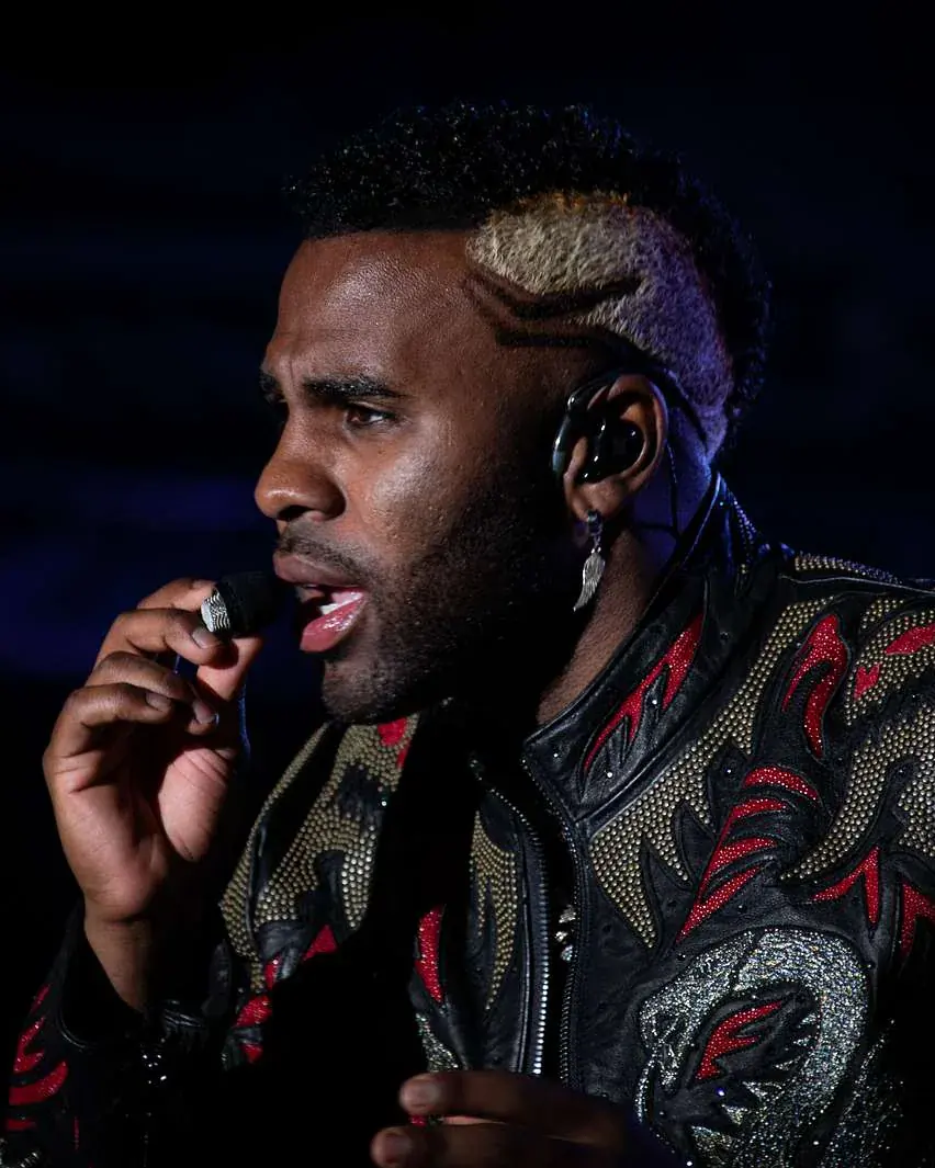Jason Derulo in profile, singing into a headset microphone, in a studded black jacket with red flame patterns