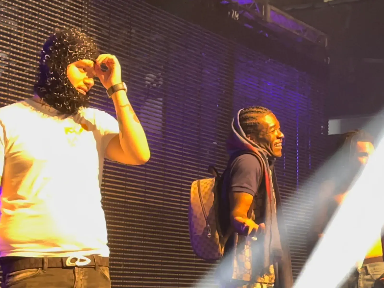 Two rappers onstage in purple light, one in a curly black balaclava and white T-shirt, the other with braids and a backpack, smiling
